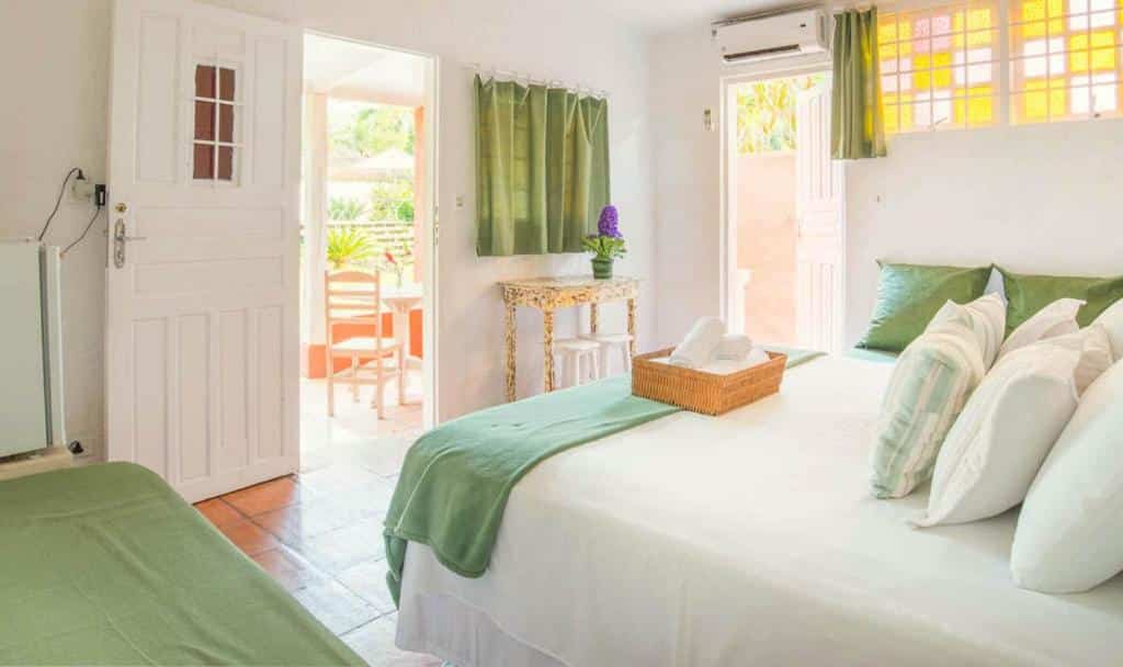 Quarto da VELINN Pousada Bromelias. No lado direito pode-se ver uma cama de casal. No lado esquerdo há uma mesinha com planta em cima e dois bancos, com janela a frente, além de uma porta que vai para a área externa do quarto. Ao fundo pode-se ver uma outra porta que vai para a fora do quarto, além de alguns vitrais. Imagem utilizada para ilustrar o post pousadas pet friendly em Ilhabela.
