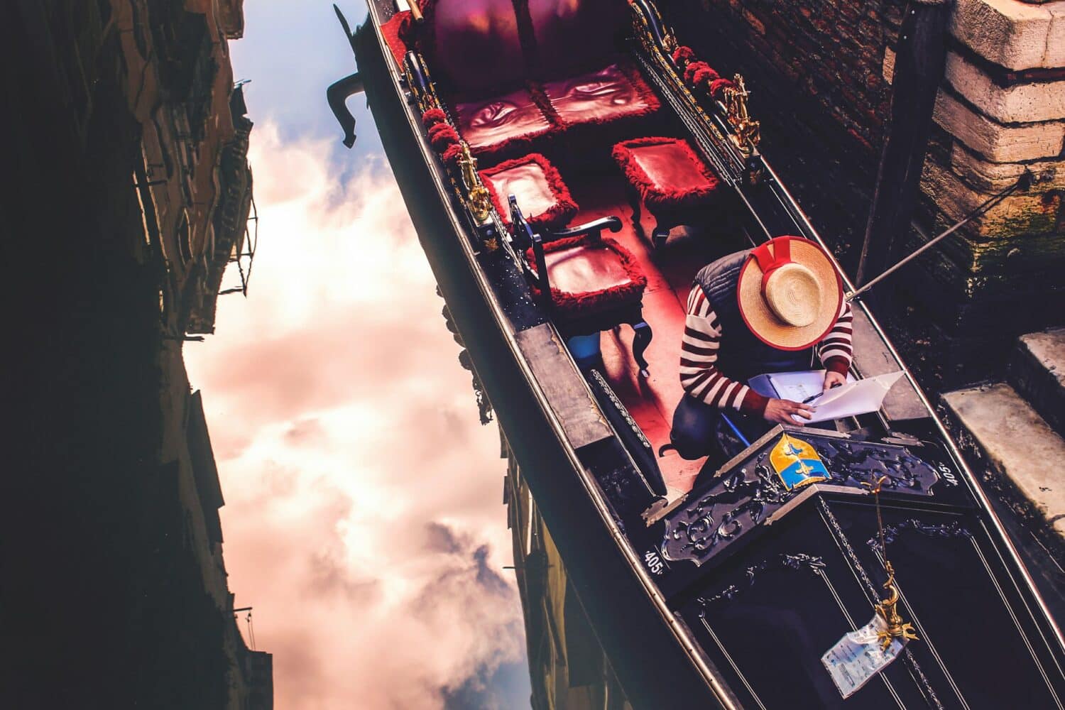 vista de cima de um barqueiro mexendo em uma pasta de documentos em cores quentes e o céu refletido nas águas dos canais de Veneza, na Itália, que pode ser visitada com o CDAM