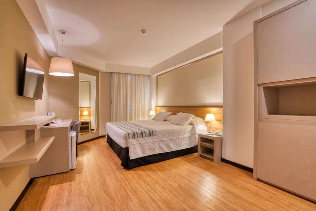 Quarto do Viale Cataratas Hotel & Eventos com uma cama de casal e do lado esquerdo dela tem uma mesa de cabeceira e um guarda-roupa. Na frente da cama tem algumas prateleiras, um frigobar, uma tv na parede e um lustre no teto. Já do lado direito da cama tem um espelho e uma cortina.