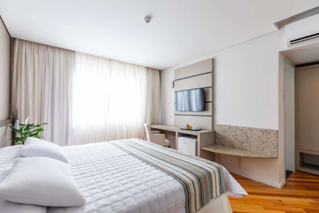 Foto do quarto do Viale Cataratas Hotel & Eventos, ilustrando o post sobre Onde ficar em Foz do Iguaçu. Há uma cama box de casal na esquerda, e na frente há uma escrivaninha com TV e frigobar. Do lado esquerdo da cama há uma janela com cortinas claras.
