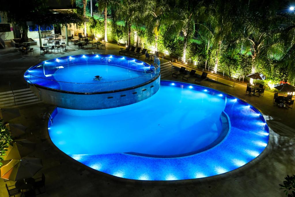 Foto do Viale Cataratas Hotel & Eventos, mostrando duas piscinas redondas e bem iluminadas à noite. Em volta do deck há cadeiras e mesas com guarda-sol e palmeiras. 