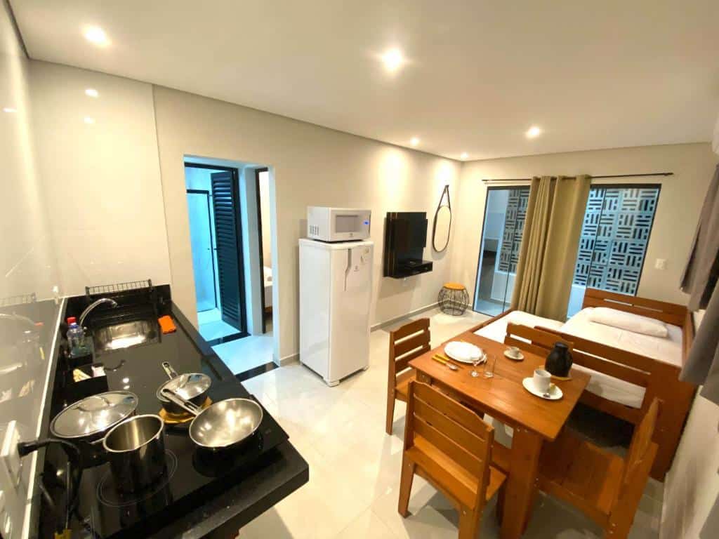 Cozinhha e quarto da Villa Licuri Boiçucanga. Do lado direito está a parte da cozinha com uma pia, fogão, mesa e geladeira. Ao fundo, do lado esquerdo, estão uma cama normal e uma no chão, de frente para uma tv na parede. Este é um bom airbnb em Boiçucanga.