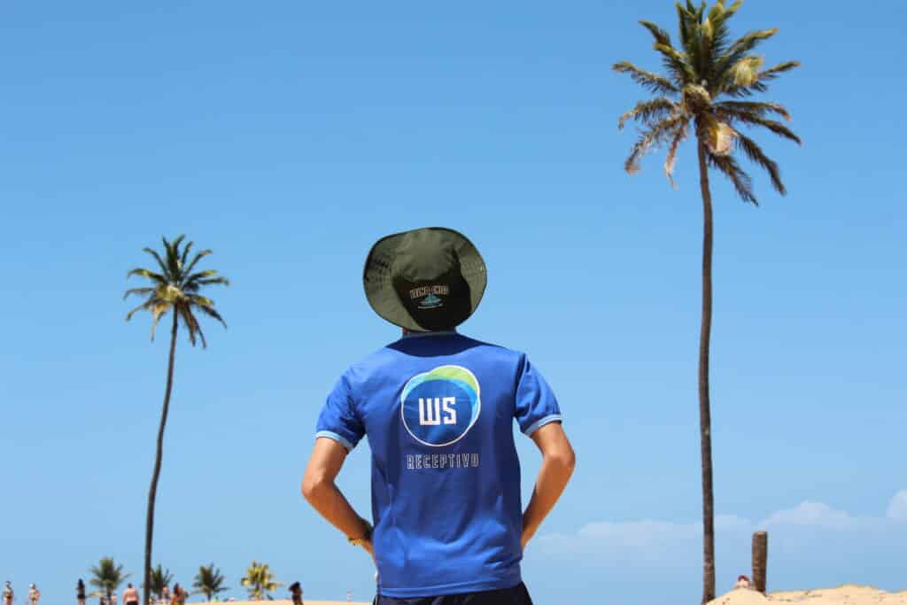 Homem de costa vestindo uma camiseta escrito "WS RECEPTIVO" e um chápeu escrito "VELHO CHICO, PIAÇABUÇU-AL". Na sua frente tem coqueiros e um céu azul sem núvens
