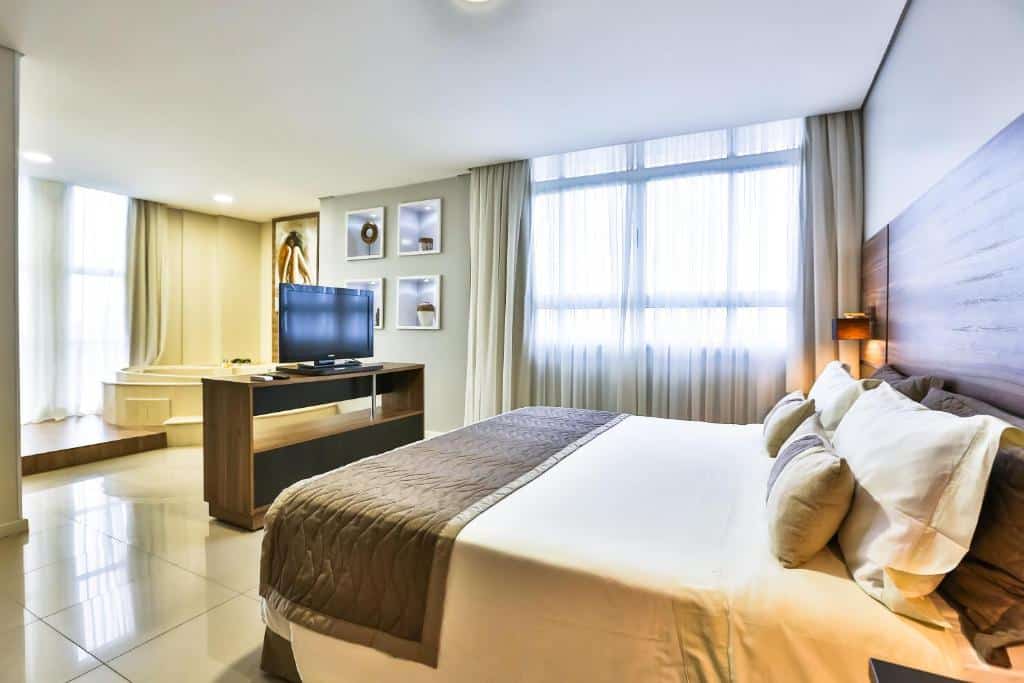 Foto do quarto do Wyndham Golden Foz Suítes, ilustrando o post sobre Onde ficar em Foz do Iguaçu. Há uma cama box de casal encostada no centro da parede à direita. Do lado dela há uma janela e na frente há uma mesa com TV.