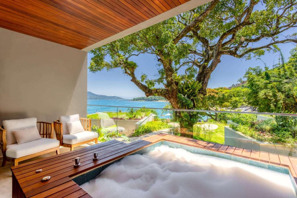 Varanda de um dos quartos do hotel Wyndham Ilhabela Casa Di Sirena. Na frente da imagem há uma banheira cheia de espuma e duas cadeiras no lado esquerdo da varanda. Já ao fundo a vista da varanda é para uma vasta vegetação a direita e para o mar a esquerda. Imagem utilizada para ilustrar o post pousadas pet friendly em Ilhabela.