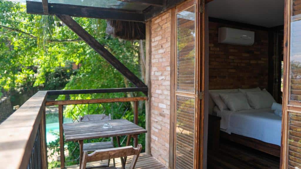 Varanda do quarto da pousada Yannai Chalé Praia. É possível ver a esquerda uma varanda com mesa e cadeiras, além de um parapeito feito em madeira e acima uma estrutura para cobrir a varanda. A direita há uma porta que vai para o quarto.