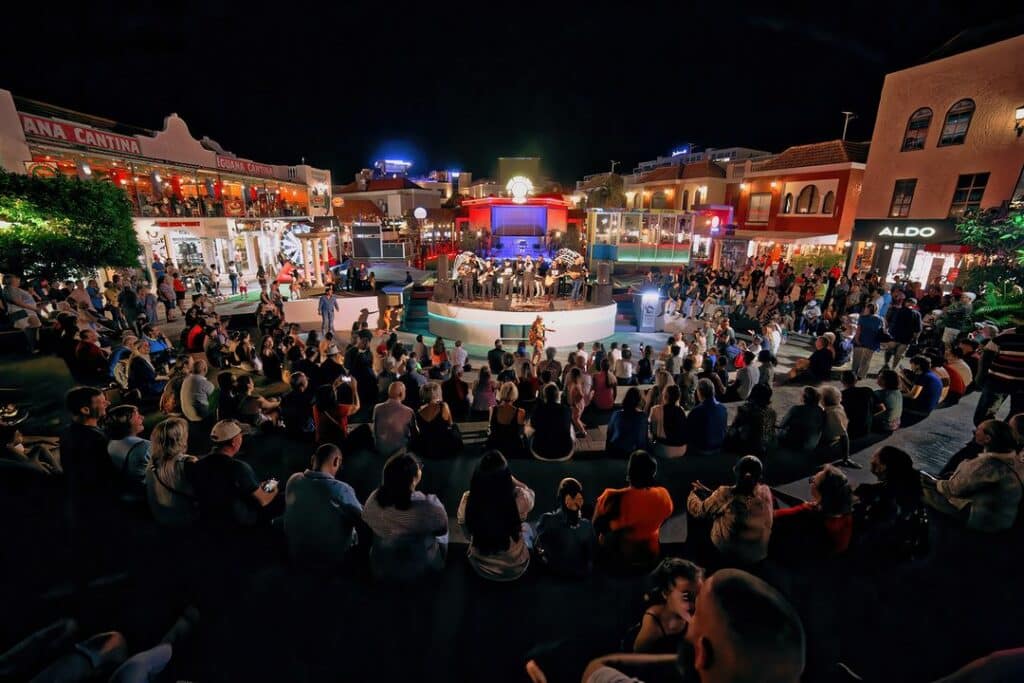 diversas pessoas sentadas em círculo ao redor de um palco principal onde está acontecendo uma apresentação musical em Paseo Herencia, em Palm Beach, um lugar para fazer compras em Aruba