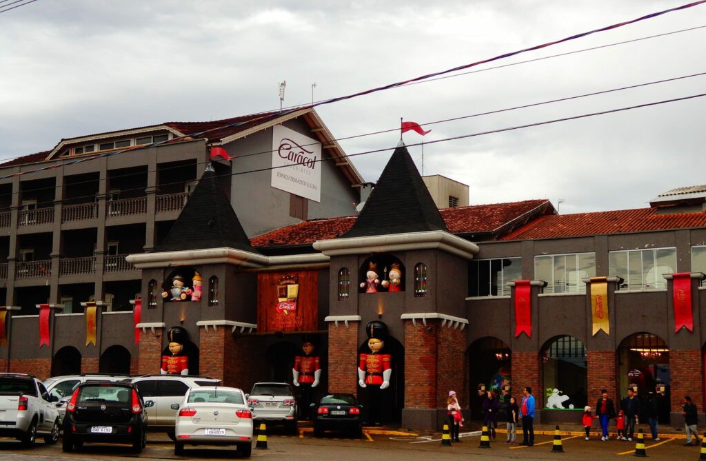 A imagem mostra a fachada do Reino do Chocolate em Gramado, RS. O edifício tem um design que lembra um castelo, com torres e decoração temática. No exterior, são visíveis várias figuras decorativas, incluindo grandes bonecos de soldadinhos de chumbo e personagens de contos de fadas. O prédio é de cor cinza com detalhes em tijolo vermelho nas bases das torres. Acima da entrada principal, há um banner indicando o nome do local: "Caracol Chocolates - Espaço Temático e Loja". No estacionamento em frente ao edifício, há vários carros estacionados e algumas pessoas caminhando e observando a fachada.
