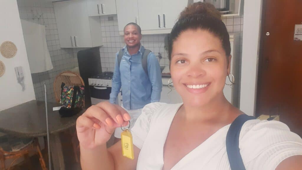 Selfie de uma mulher vestida de branco segurando uma chave com uma mão um homem atrás sorrindo com mochila nas costas. Eles estão dentro de um apartamento, com a pia da cozinha e mesa atrás. Representa Airbnb é confiável