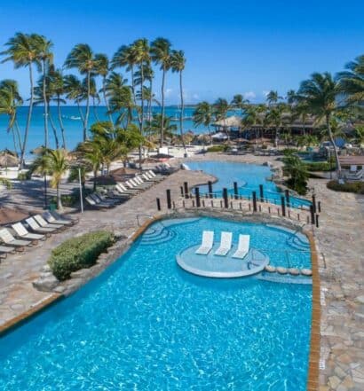 Área externa do All Inclusive Holiday Inn Resort Aruba. Duas piscinas no meio, do lado esquerdo cadeiras de tomar sol e palmeiras, atrás o mar. Foto para ilustrar post sobre hotéis all inclusive em Aruba.