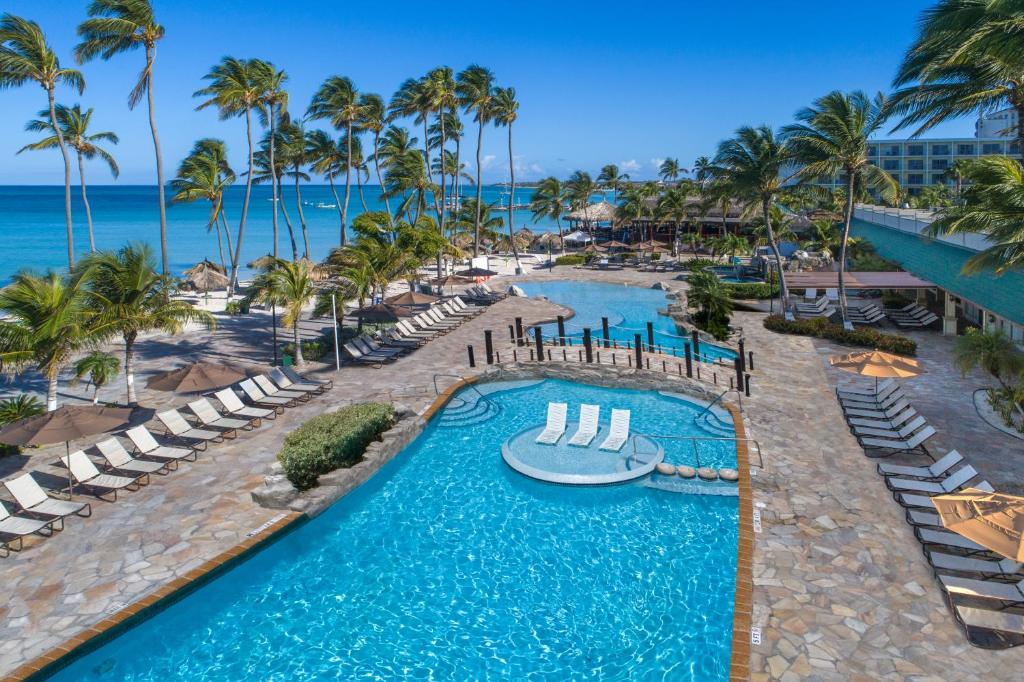 Área externa do All Inclusive Holiday Inn Resort Aruba. Duas piscinas no meio, do lado esquerdo cadeiras de tomar sol e palmeiras, atrás o mar. Foto para ilustrar post sobre hotéis all inclusive em Aruba.