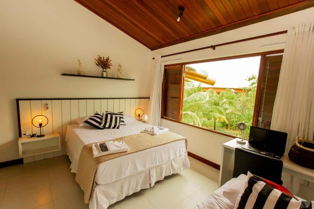 Quarto do Apartamento dos Sonho Imbassaí. Há uma cama de casal no centro, e do lado direito dela há uma janela com vista. Há uma prateleira em cima da cama, na parede, com algumas decorações. Na direita, no canto, há uma mesa de escrivaninha com computador e cadeira.