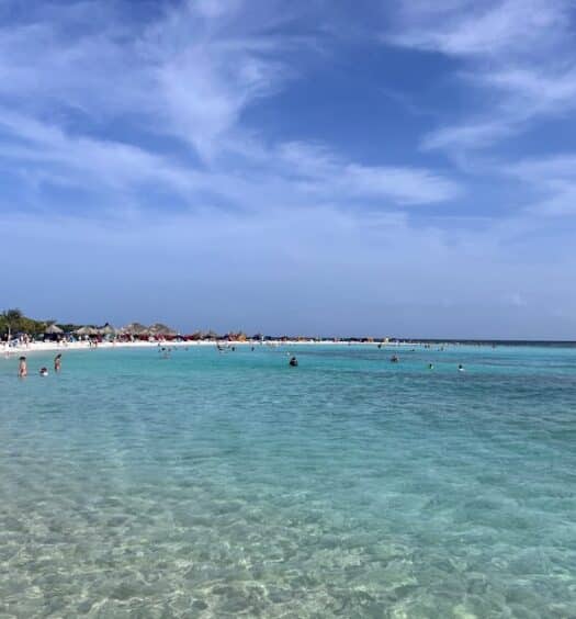 Imagem de Baby Beach para ilustrar post sobre como chegar em Aruba. Um mar cristalino sem ondas, no fundo algumas pessoas na água.