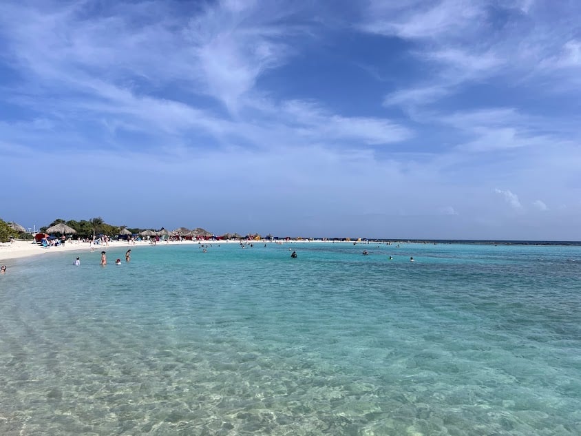 Imagem de Baby Beach em Aruba. Um mar cristalino sem ondas, no fundo algumas pessoas na água.