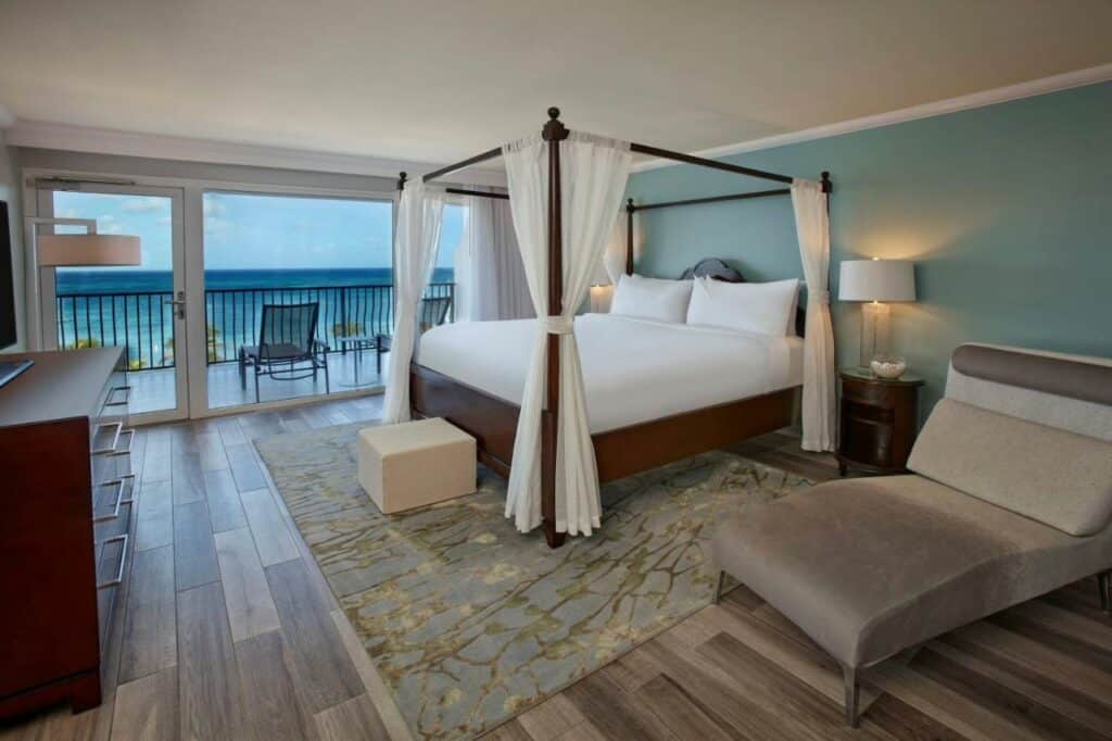 Quarto do Aruba Marriott Resort & Stellaris Casino. Uma cama de casal com dossel está no meio, ao seu lado direito um recamier. Ao fundo está a varanda, que possui algumas espreguiçadeiras e vista para o mar. Ele faz parte dos hotéis de luxo em Aruba.