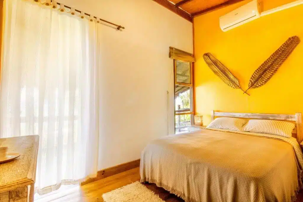 Quarto do Bangalô Ilhabela. Uma cama de casal está encostada na parede ao lado direito. Uma decoração de parede fica logo acima, assim como um aparelho de ar-condicionado. Em frente à cama está uma cômoda de madeira, e ao lado esquerdo a parede tem duas janelas com cortinas.