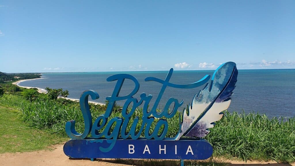 Um placa presa no chão escrito Porto Seguro e, ao fundo, há o mar azul e a orla de uma praia