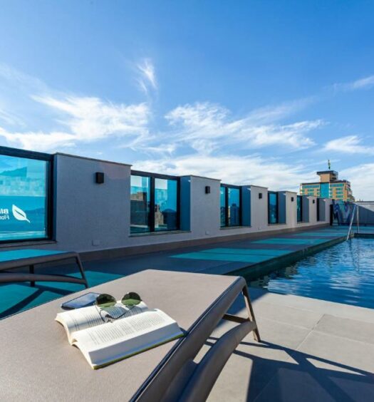 Vista do terraço no Blue Tree Premium Florianópolis Há grades de vidro. Uma piscina e um deck com espreguiçadeira, champagne na beira e um livro com óculos de sol em cima da espreguiçadeira.