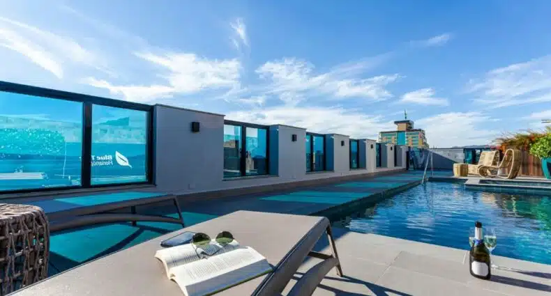 Vista do terraço no Blue Tree Premium Florianópolis Há grades de vidro. Uma piscina e um deck com espreguiçadeira, champagne na beira e um livro com óculos de sol em cima da espreguiçadeira.