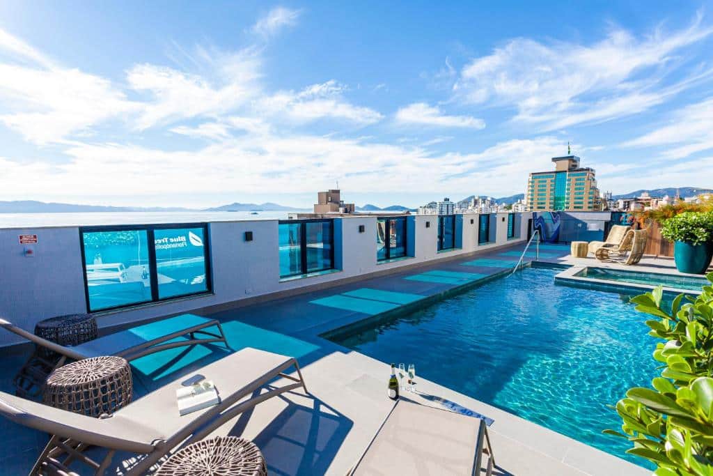 Vista do terraço no Blue Tree Premium Florianópolis Há grades de vidro. Uma piscina e um deck com espreguiçadeiras, champagne na beira e um livro com óculos de sol em cima de uma das espreguiçadeiras.