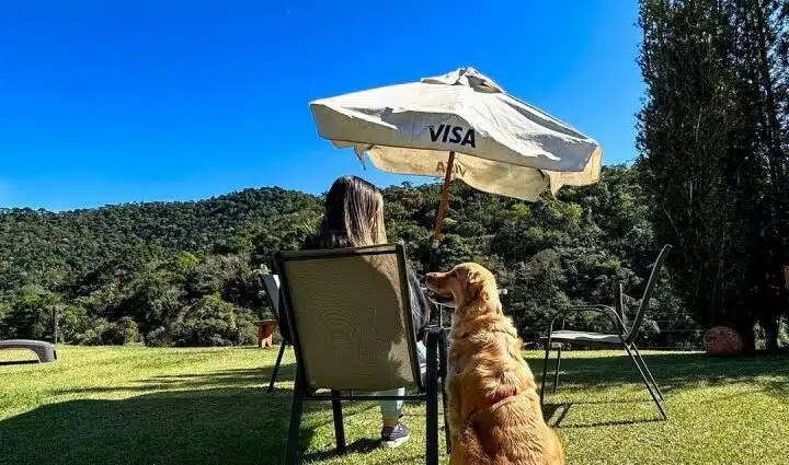 Cachorro ao lado de uma pesso sentada em cadeira e debaixo de uma espreguiçadeiras. Os dois estão em um gramado e de frente para uma área com vegetação. Essa é uma imagem para o post de pousadas pet friendly em Santo Antônio do Pinhal.