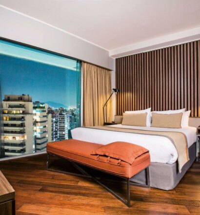Quarto do DoubleTree by Hilton Santiago Kennedy com cama de casal no centro do quarto no pé da cama uma poltrona e do lado direito da cama uma janela ampla com vista para a cidade. Representa hotéis 4 estrelas em Santigo.