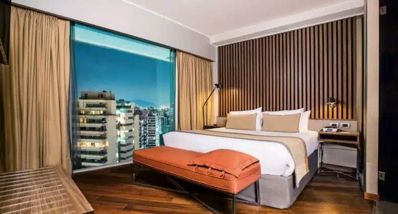 Quarto do DoubleTree by Hilton Santiago Kennedy com cama de casal no centro do quarto no pé da cama uma poltrona e do lado direito da cama uma janela ampla com vista para a cidade. Representa hotéis 4 estrelas em Santigo.