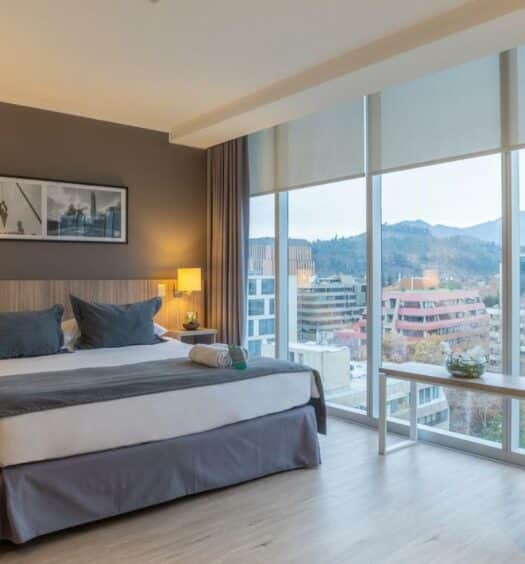 Quarto do Hotel Capital Bellet com cama de casal do lado esquerdo da imagem com uma comoda do lado esquerdo da cama e do lado esquerdo um janela ampla com vista para a cidade. Representa hotéis 3 estrelas em Santiago.