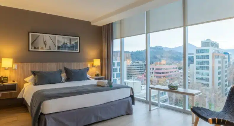 Quarto moderno do Hotel Capital Bellet com uma cama grande, duas mesinhas de cabeceira e janelas do chão ao teto com vista para os prédios da cidade e as montanhas. Representa hotéis 3 estrelas em Santiago.