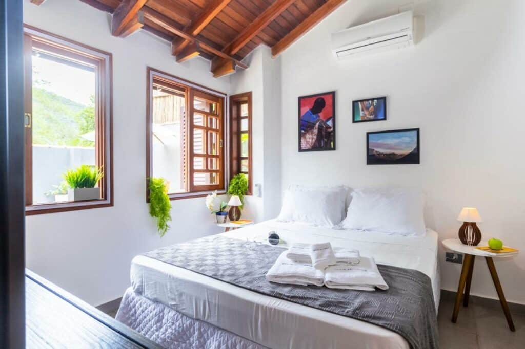 Quarto da Casa Charmosa com Piscina em Barra do Una, um dos airbnb em Barra do Una. Uma cama de casal tem toalhas aos pés e mesinhas com abajures dos dois lados. Na parede acima há quadrinhos e um aparelho de ar-condicionado, e ao lado esquerdo ficam duas janelas abertas.