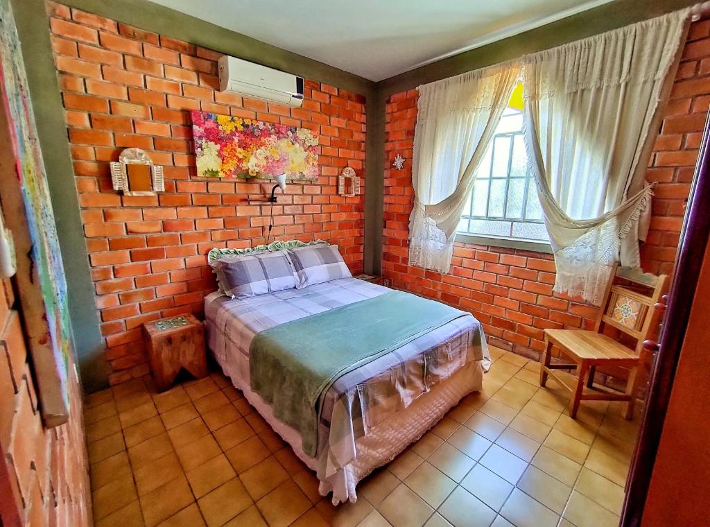 Um pequeno quarto com paredes de tijolos, uma cama de casal, móveis de madeira, uma janela com cortinas e um ar-condicionado, na Casa Rústica em Florianópolis.