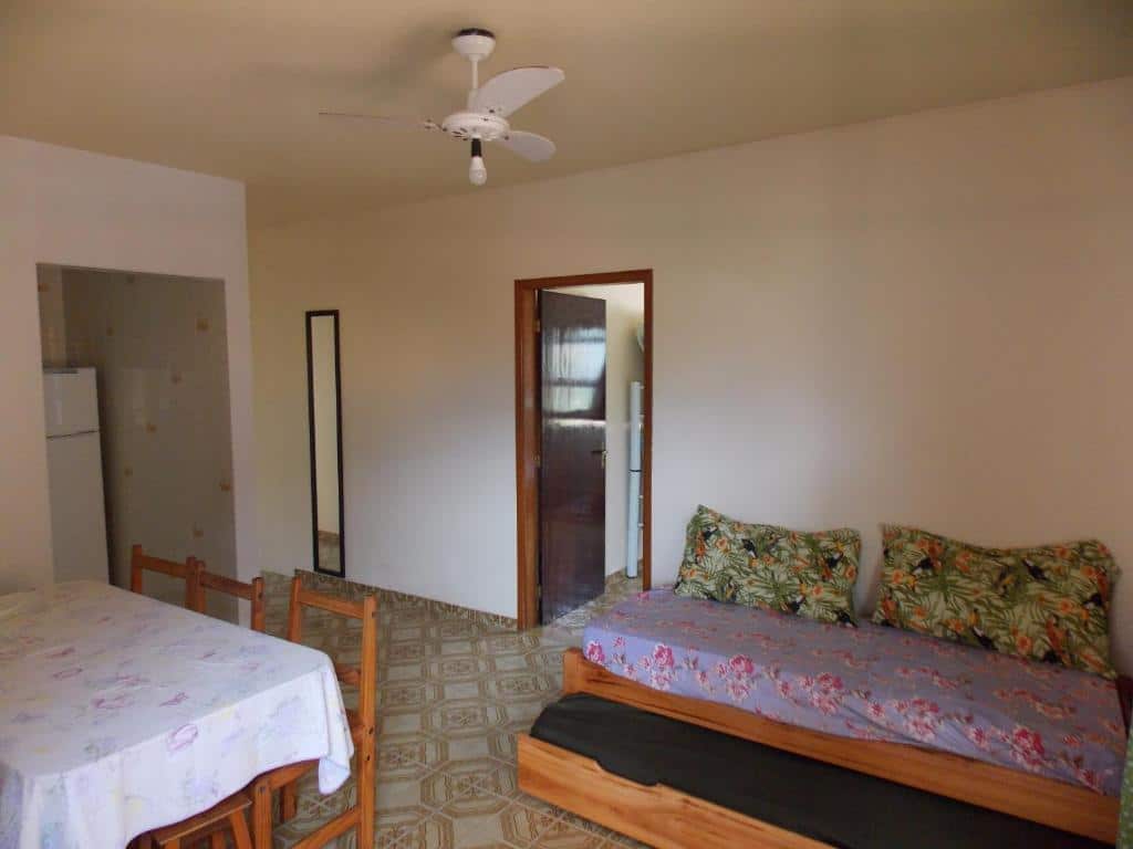 Foto de um quarto com um sofá-cama na direita, uma mesa de jantar com quatro cadeiras e uma toalha floral na esquerda, um ventilador de teto branco, um espelho de corpo inteiro e uma porta aberta que leva a outro cômodo. Ilustra o post de pousada em Maranduba.