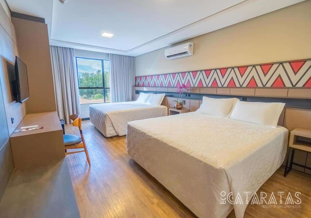 Quarto do Eco Cataratas Resort by San Juan que mostra duas camas de casal lado a lado e de frente um painel com tv e uma escrivaninha. No quarto também tem um ar-condicionado e uma janela de vidro com cortinas.