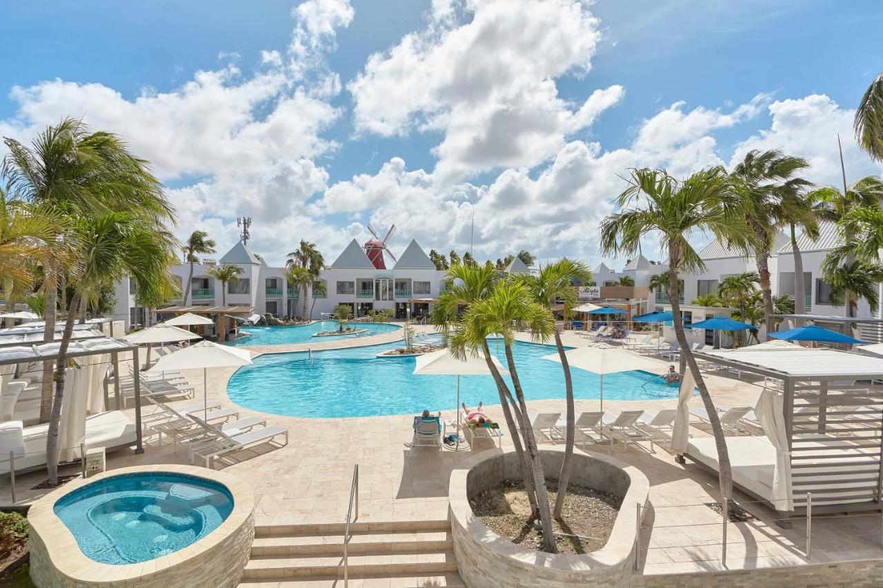 Piscinas do Courtyard by Marriott Aruba Resort. Elas estão lado a lado em uma área aberta, cercada por guarda-sóis, espreguiçadeiras e árvores.