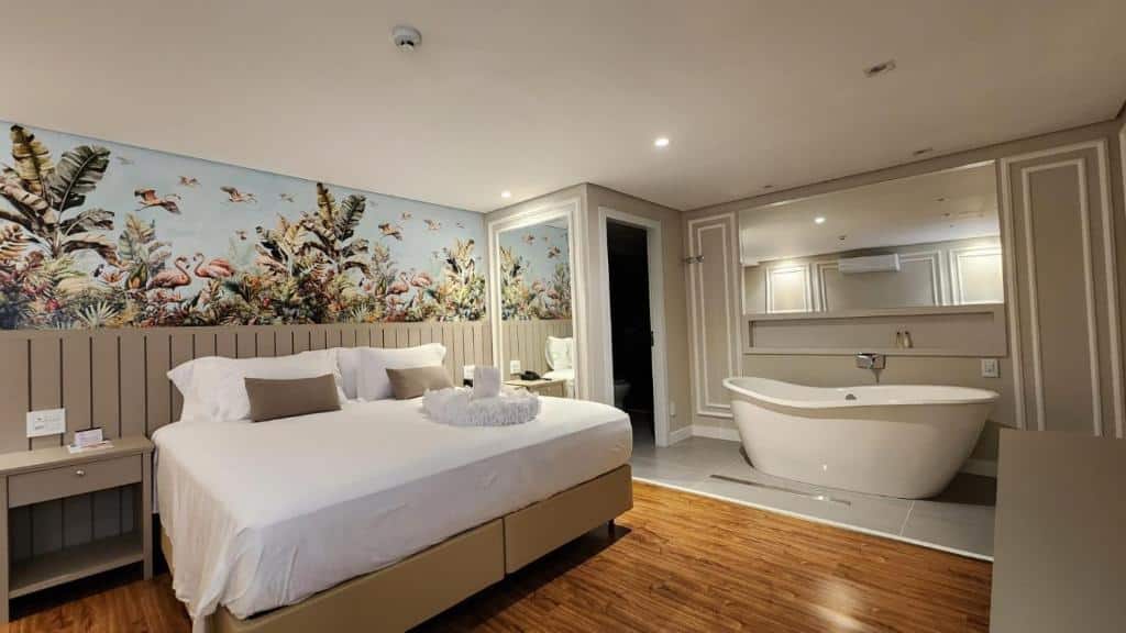 Foto do quarto do Del Rey Quality Hotel, ilustrando o post sobre Onde ficar em Foz do Iguaçu. Há uma cama box de casal no centro, atrás há uma parede com papel de parede tropical. Na direita da cama há um espelho refletindo a mesma. E mais para a direita do quarto, há uma banheira com outro espelho atrás.