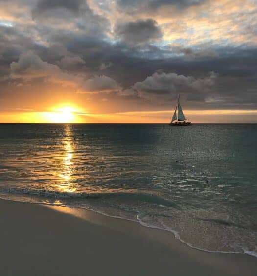 Imagem da praia de Eagle Beach, no final da tarde com faixa de areia do lado esquerdo e do lado direito o mar com um barco navegando. Representa praias em Aruba.
