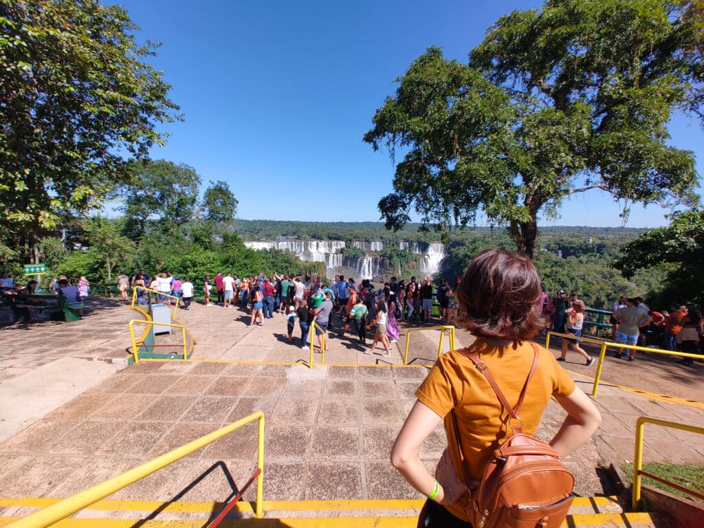 Uma foto tirada na entrada do Parque Nacional do Iguaçu. Há uma mulher virada de costas, posicionada mais à direita da foto, com as mãos na cintura olhando a vista. Ela tem cabelos curtos acastanhados, usa uma blusa laranja e uma mochila de couro. No fundo há várias pessoas observando as Cataratas do Iguaçu.