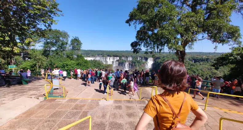 mulher de costas olhando para as cataratas do iguaçu à sua frente. Há várias pessoas aglomeradas próximas ao guarda-corpo em frente as cataratas, onde estão tirando fotos e admirando a paisagem