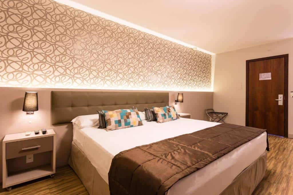 Foto do quarto do hotel Faial Prime Suítes. Ilustra o post sobre Onde ficar em Florianópolis. Há uma cama box de casal no centro. Atrás dela há uma cabeceira e luminárias. Dos dois lados da cama há uma mesa de cabeceira com gaveta. Na direita há uma porta.