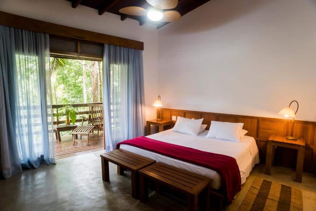 Foto do quarto em Guest House Ilha Splendor. Ilustra o post sobre Onde ficar em Ilhabela. O quarto é espaçoso, há uma cama box de casal no centro e de seus dois lados há uma mesa de cabeceira com abajur. No lado esquerdo do quarto há uma porta grande com varanda e vista.