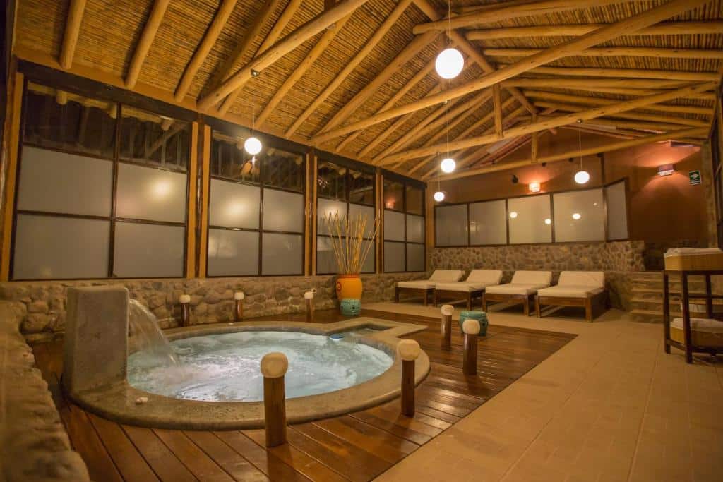Área de jacuzzi do Tierra Viva Valle Sagrado Hotel. O espaço é fechado e todo trabalhado com pedras e teto de madeira. Há quatro espreguiçadeiras brancas ao lado da jacuzzi.