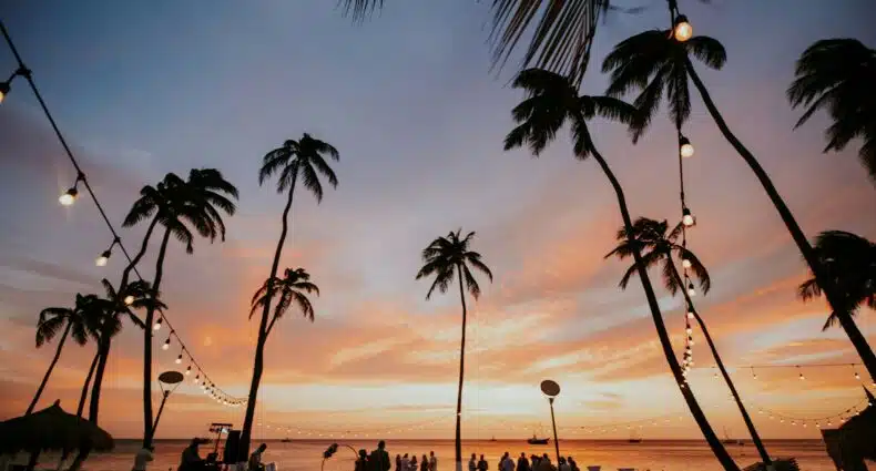 palmeiras rasgando o céu alaranjado do Hilton Aruba Caribbean Resort & Casino, com pessoas ao redor de mesas e um DJ começando a tocar para agitar a vida noturna em Aruba