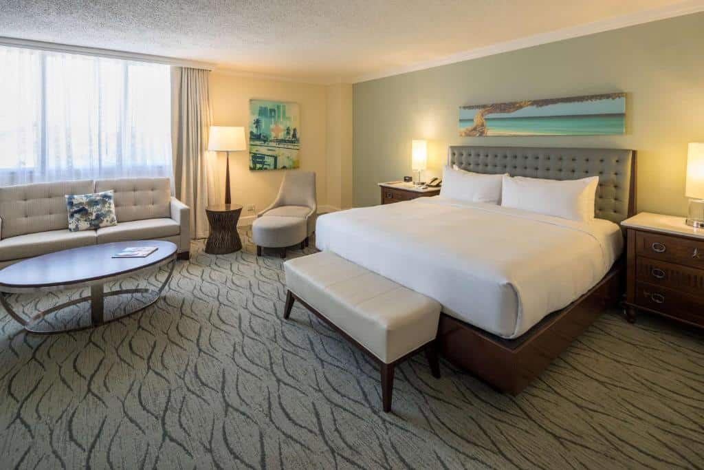 Quarto do Hilton Aruba Caribbean Resort & Casino. Uma cama de casal está no centro, um banco estofado em sua ponta. Ao seu lado esquerdo, encostado na parede com janela está um sofá, um abajur e uma poltrona. Em frente ao sofá há uma mesa redonda de centro. O chão é todo de carpete.