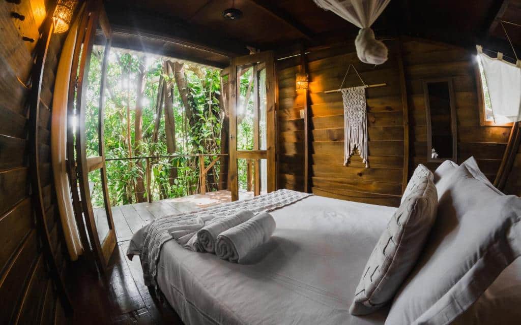 Foto do quarto em Hostel Da Vila Ilhabela. Ilustra o post sobre Onde ficar em Ilhabela. Há uma cama box de casal na direita, e na frente dela há uma porta extensa com varanda e vista. As paredes são de madeira e dão um ar rústico.