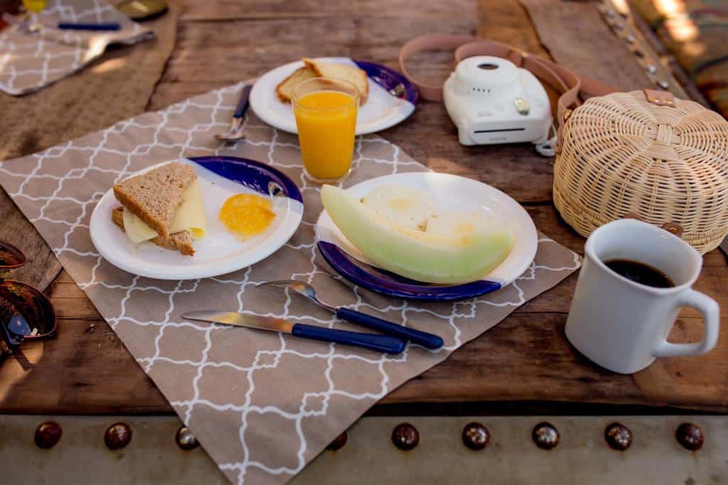 Mesa mostrando um café da manhã no Hostel Da Vila Ilhabela. Há um prato com fruta, pão integral, um copo com suco e uma xícara com café Também há uma câmera polaroid sobre a mesa.