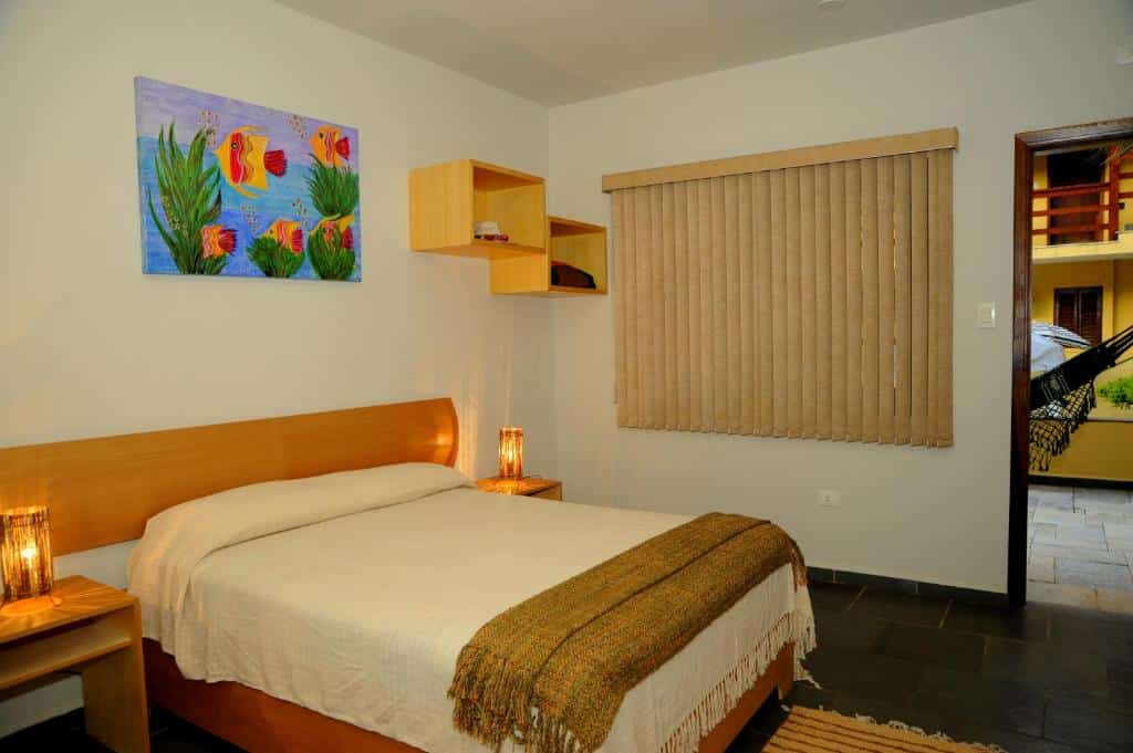 Foto do quarto em Hotel da Ilha. Ilustra o post sobre Onde ficar em Ilhabela. Há uma cama box de casal na esquerda. Na direita da cama há uma janela com cortinas e porta.