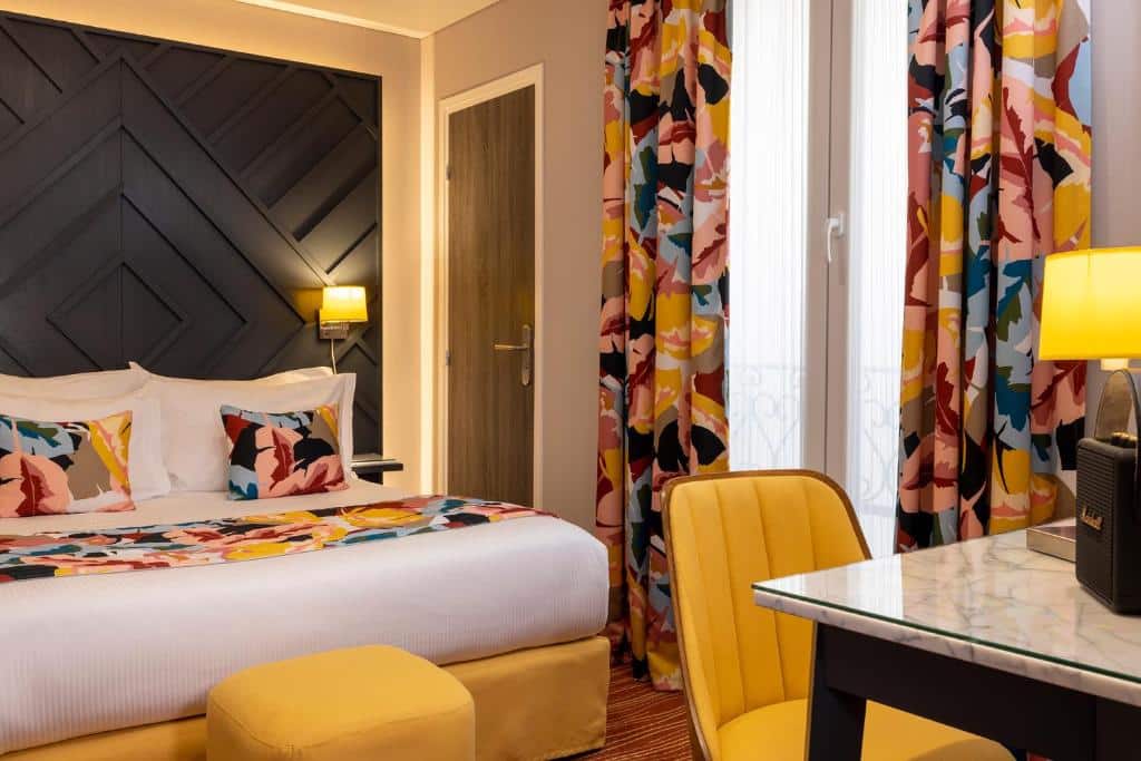 Quarto do Hôtel des Arts Montmartre, um dos hotéis 4 estrelas em Paris. Uma cama de casal está encostada na parede ao lado esquerdo com mesinhas de cabeceira e luminárias aos dois lados, assim como um banco aos pés. Ao lado fica uma janela com cortinas, e em frente à cama há uma mesa com cadeira e abajur.