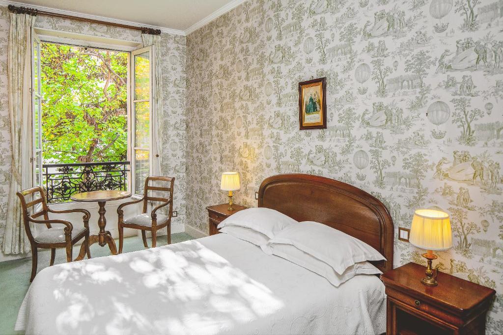 Quarto do Hotel des Grandes Ecoles, um dos hotéis 3 estrelas em Paris. Uma cama de casal está ao lado direito da imagem com mesinhas de cabeceira com luminárias dos dois lados. Na lateral esquerda da cama fica uma porta de vidro com cortinas e vista para árvores no jardim, e logo em frente há uma mesinha de madeira com duas cadeiras.
