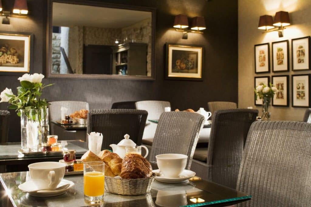 Sala de refeições do Hotel du Champ de Mars. As mesas e cadeiras estão dispostas por ali com itens de café da manhã, como pães, sucos e xícaras de café. Ao fundo é possível ver um espelho e luminárias acima dos quadrinhos na parede.