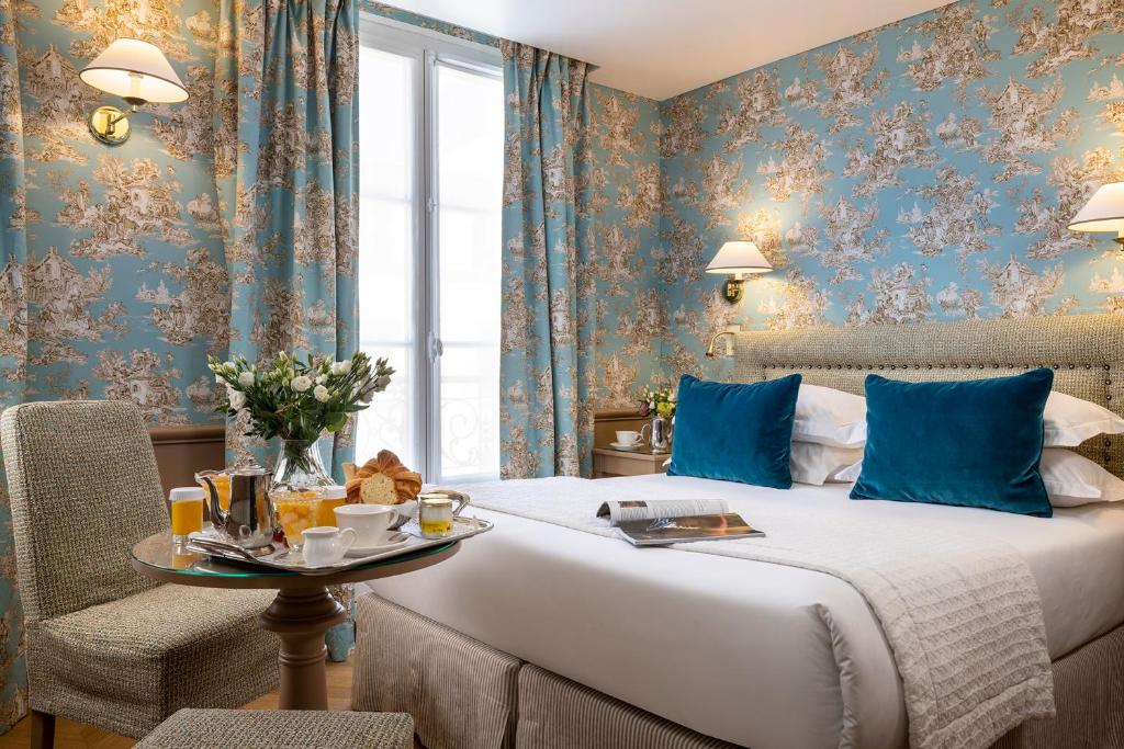 Quarto do Hotel du Champ de Mars, um dos hotéis 3 estrelas em Paris. Uma cama de casal está ao lado direito do local com mesinhas de cabeceira e luminárias dos dois lados. Em sua frente há uma mesinha com cadeira e vários itens de café da manhã. Ao lado esquerdo da cama há uma janela com cortinas da mesma estampa do papel de parede florido.